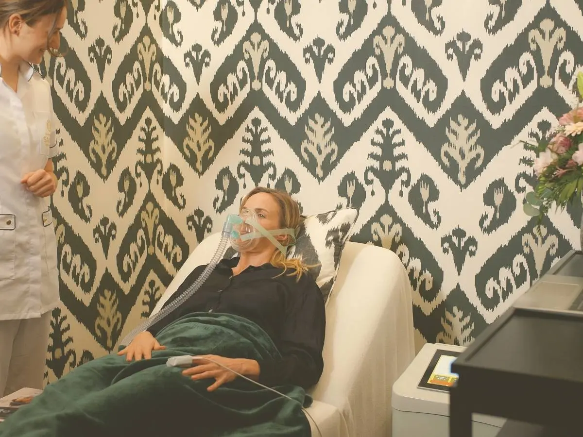 A woman lies in a treatment chair wearing a breathing mask and a finger sensor during an oxygen therapy session at the Sacher Academy for Better Aging