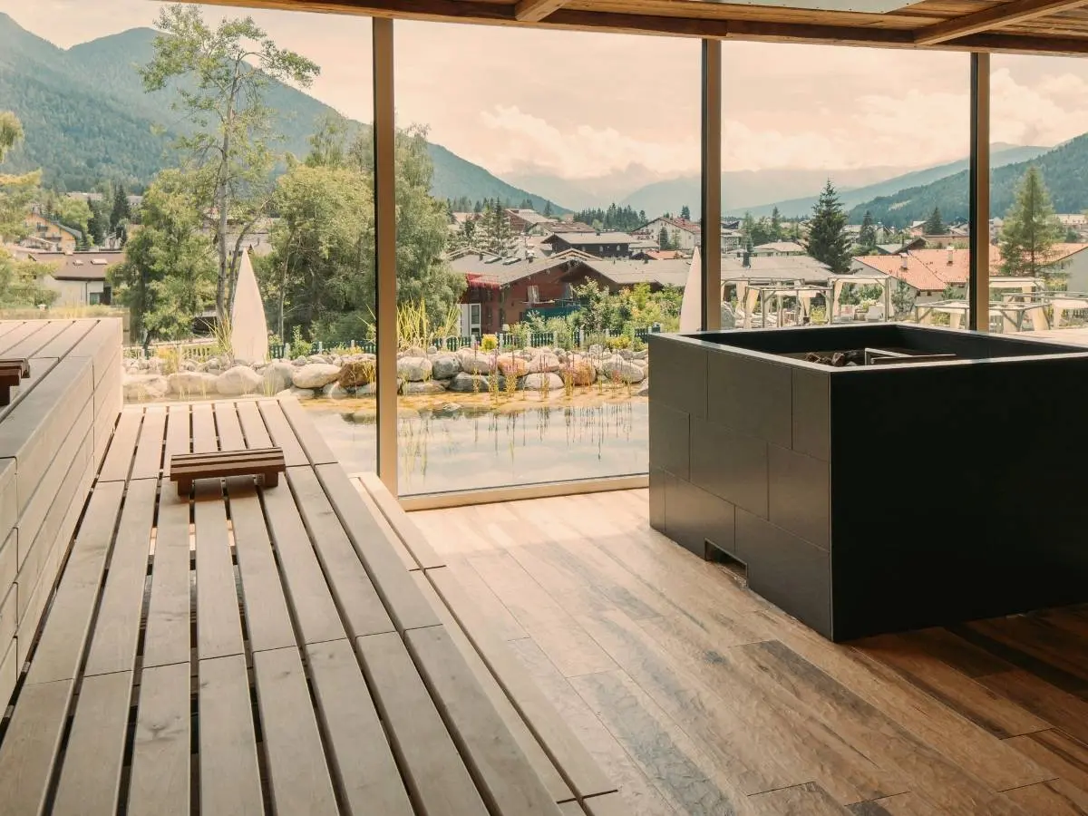 Panoramic sauna with wooden benches and a sauna stove at the Alpin Resort Sacher Seefeld, overlooking a natural pond and the Tyrolean mountains